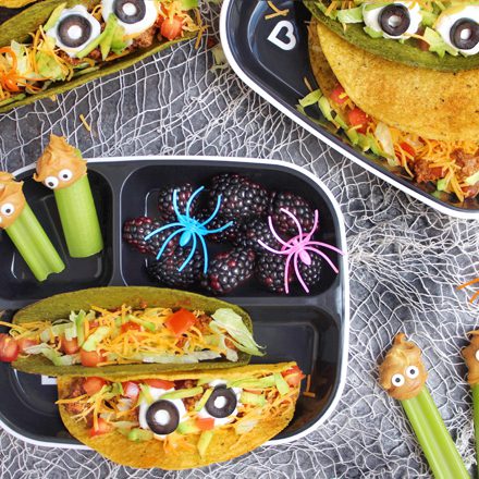 Halloween-themed children's meal trays with tacos decorated with googly eyes, celery sticks with googly eye toppers, and compartments containing berries and spider decorations on a white spiderweb background.