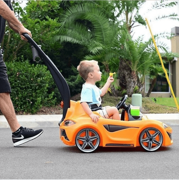 Toddler riding in an orange push-car toy while an adult holds the handle, in a driveway with tropical plants in the background.