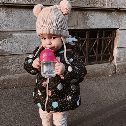 Toddler wearing a beige knit hat with ears holding a pink Munchkin sippy cup outdoors.
