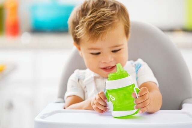 Toddler holding and drinking from a green and white Munchkin sippy cup while seated in a high chair.
