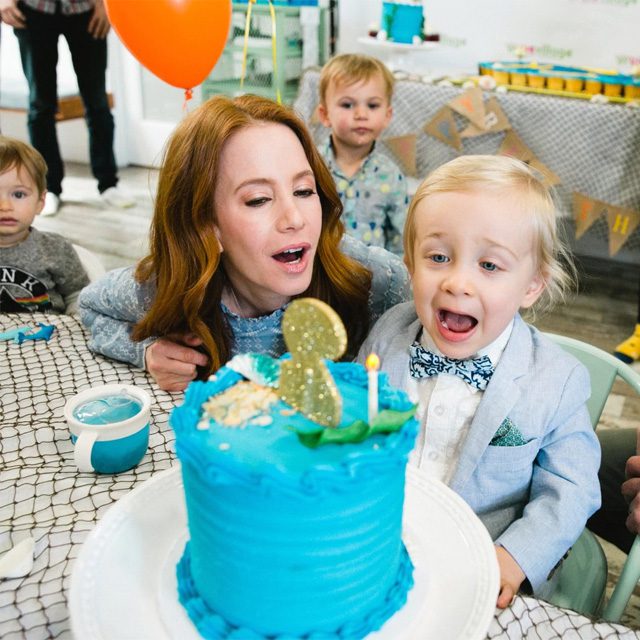 Woman and toddler celebrating with a blue cactus-themed birthday cake and orange balloon at a children's party.