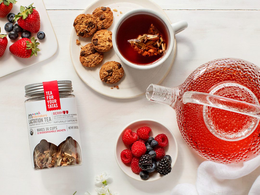 Munchkin Milkmakers Lactation Tea with berry flavor displayed alongside fresh berries, cookies, brewed tea, and a red textured teapot on a white wooden surface.