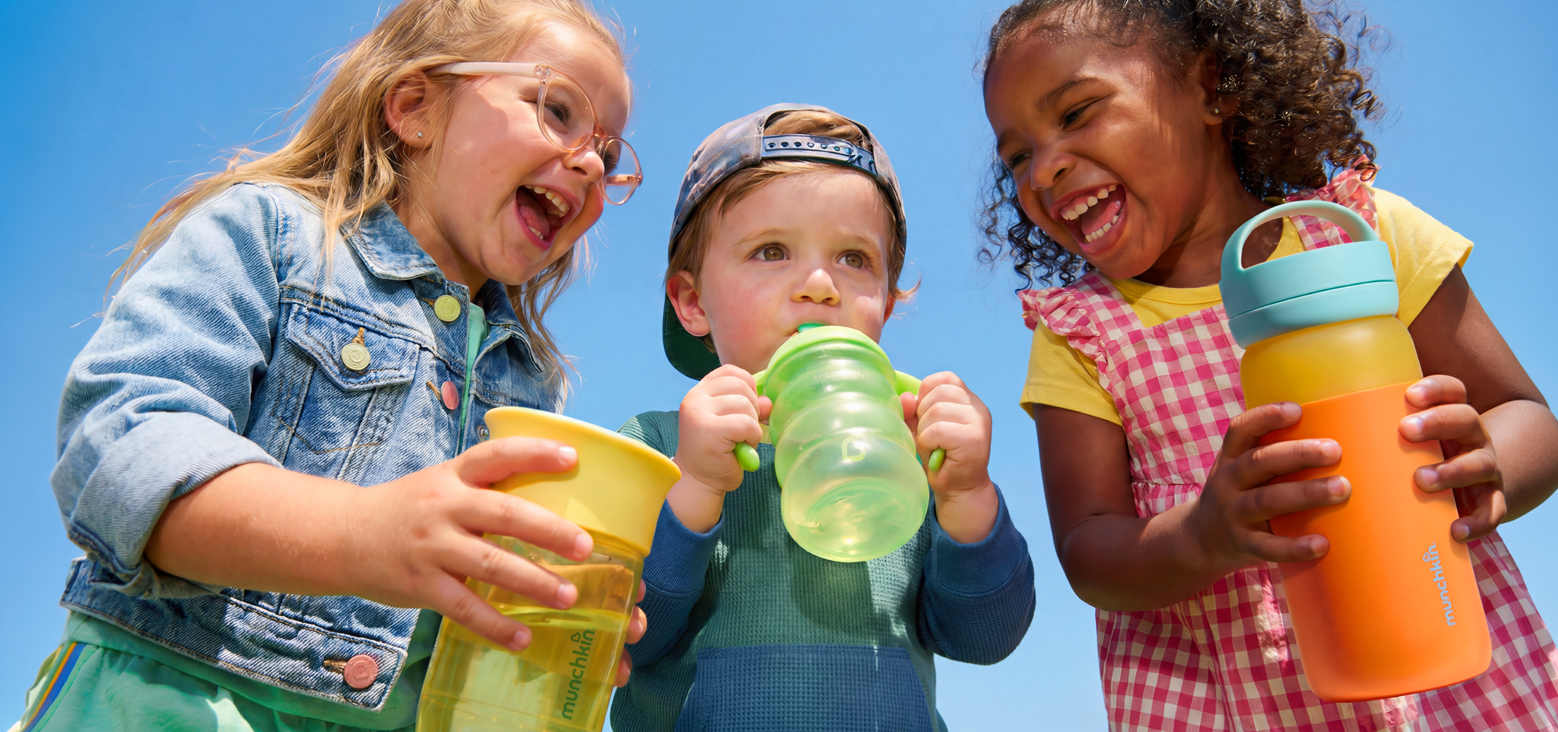 Three children holding colorful Munchkin sippy cups and bottles against a blue sky, smiling and playing outdoors.
