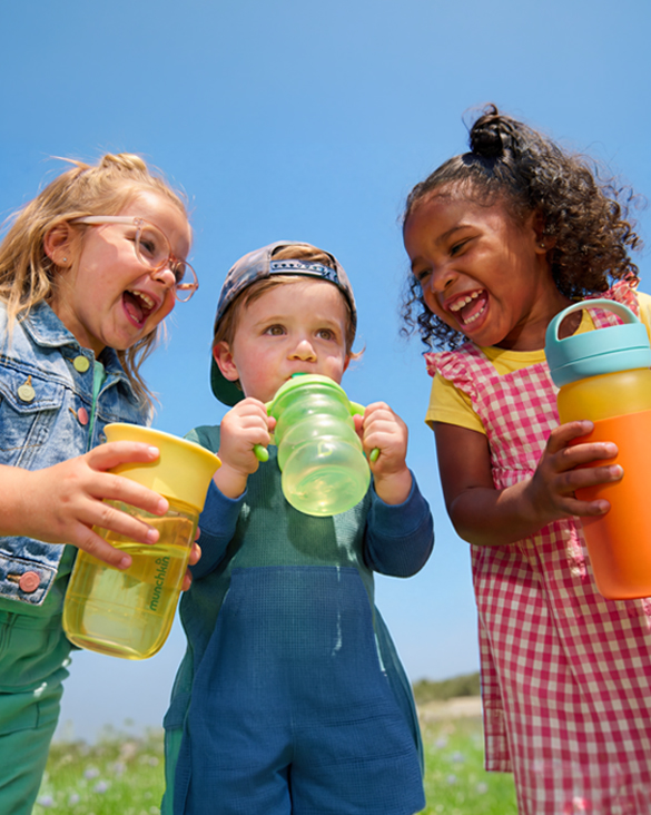 Three happy children outdoors holding colorful Munchkin sippy cups and bottles against a bright blue sky.