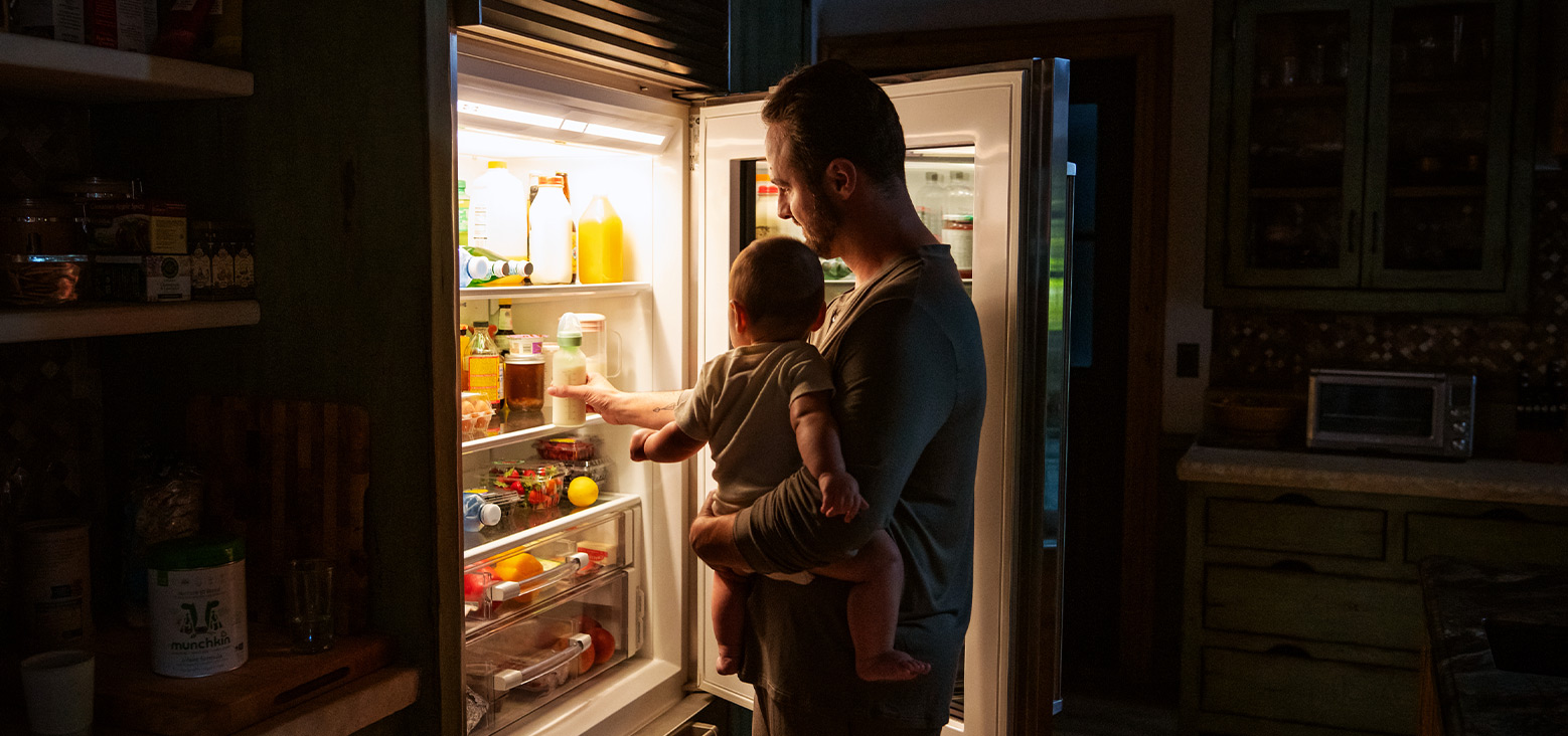 Parent holding toddler while opening a refrigerator in a kitchen at night