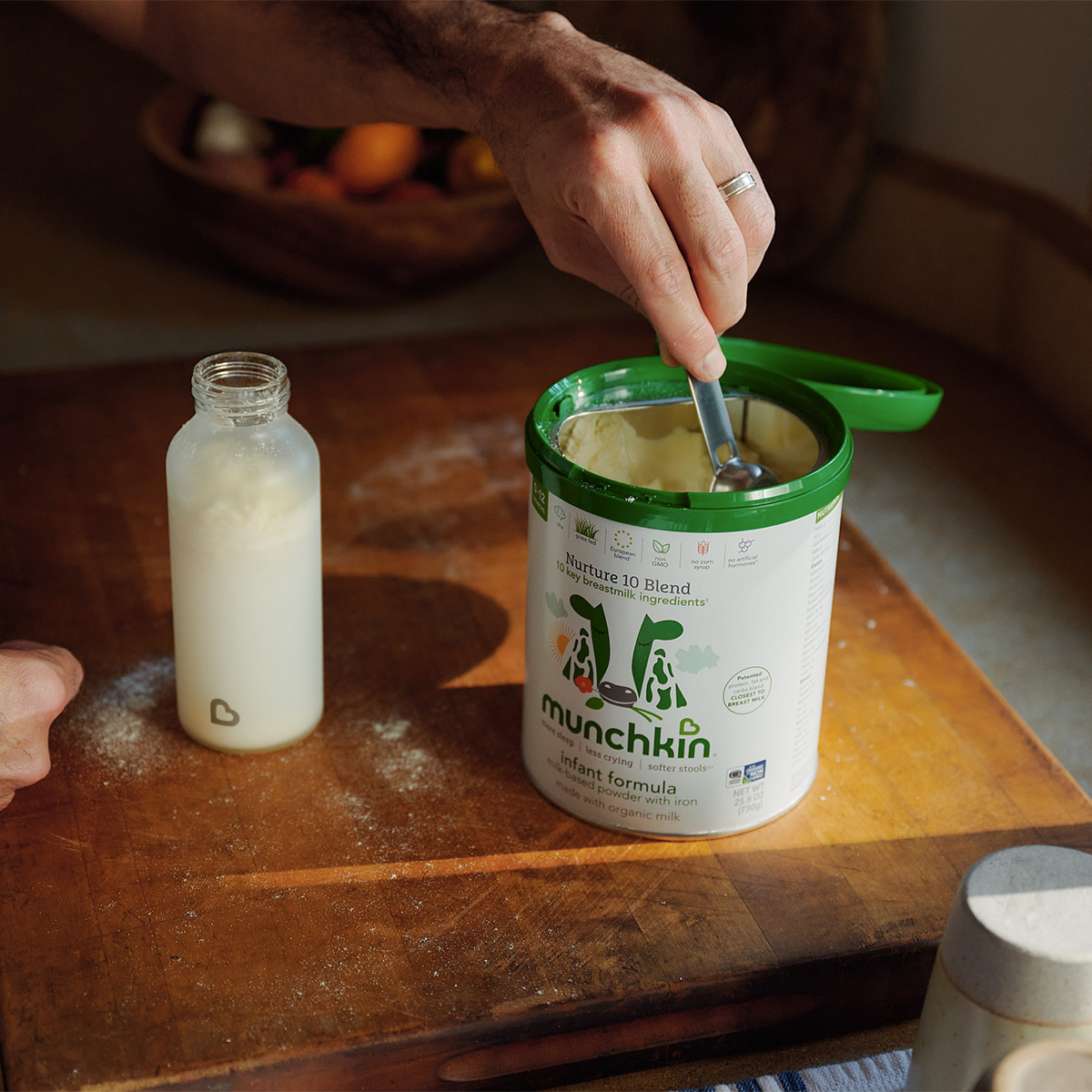 Adult preparing Munchkin Nurture 10 Blend infant formula with a scoop next to a bottle of prepared formula on a wooden surface.