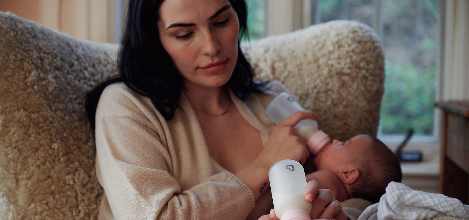 Parent holding a baby while using a Munchkin bottle warmer on a couch at home.