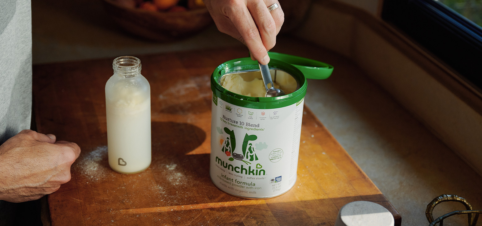 Adult scooping Munchkin Nurture 10 Blend infant formula powder from a container with a green lid, with a prepared bottle of formula beside it on a wooden table.