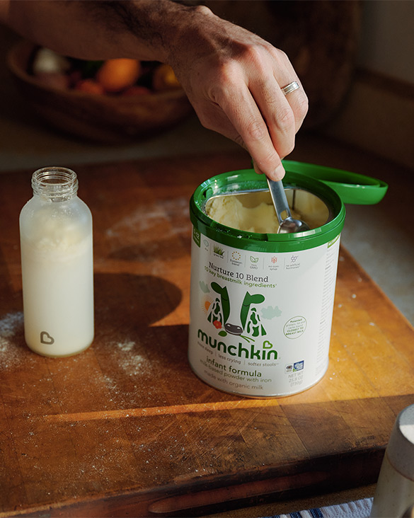 Adult hand scooping Munchkin Nature Blend infant formula from a green container, with a bottle of prepared formula nearby on a wooden surface.