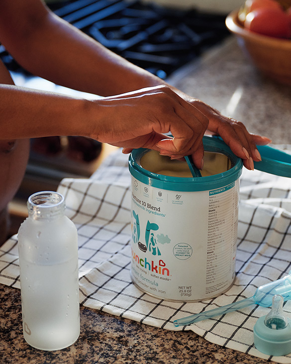Child's hands opening a teal Munchkin infant formula container next to a bottle on a checkered cloth.