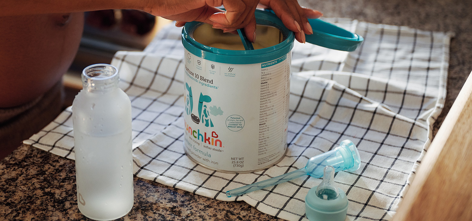 Parent opening a teal-lidded container of Munchkin infant formula on a kitchen counter, with a bottle and feeding accessories nearby.