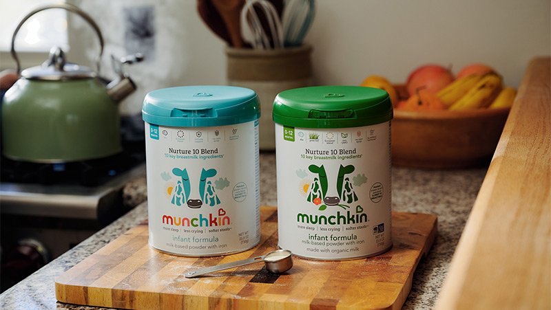 Two Munchkin infant formula containers with turquoise and green lids on a wooden cutting board in a kitchen, with a kettle and produce visible in the background.