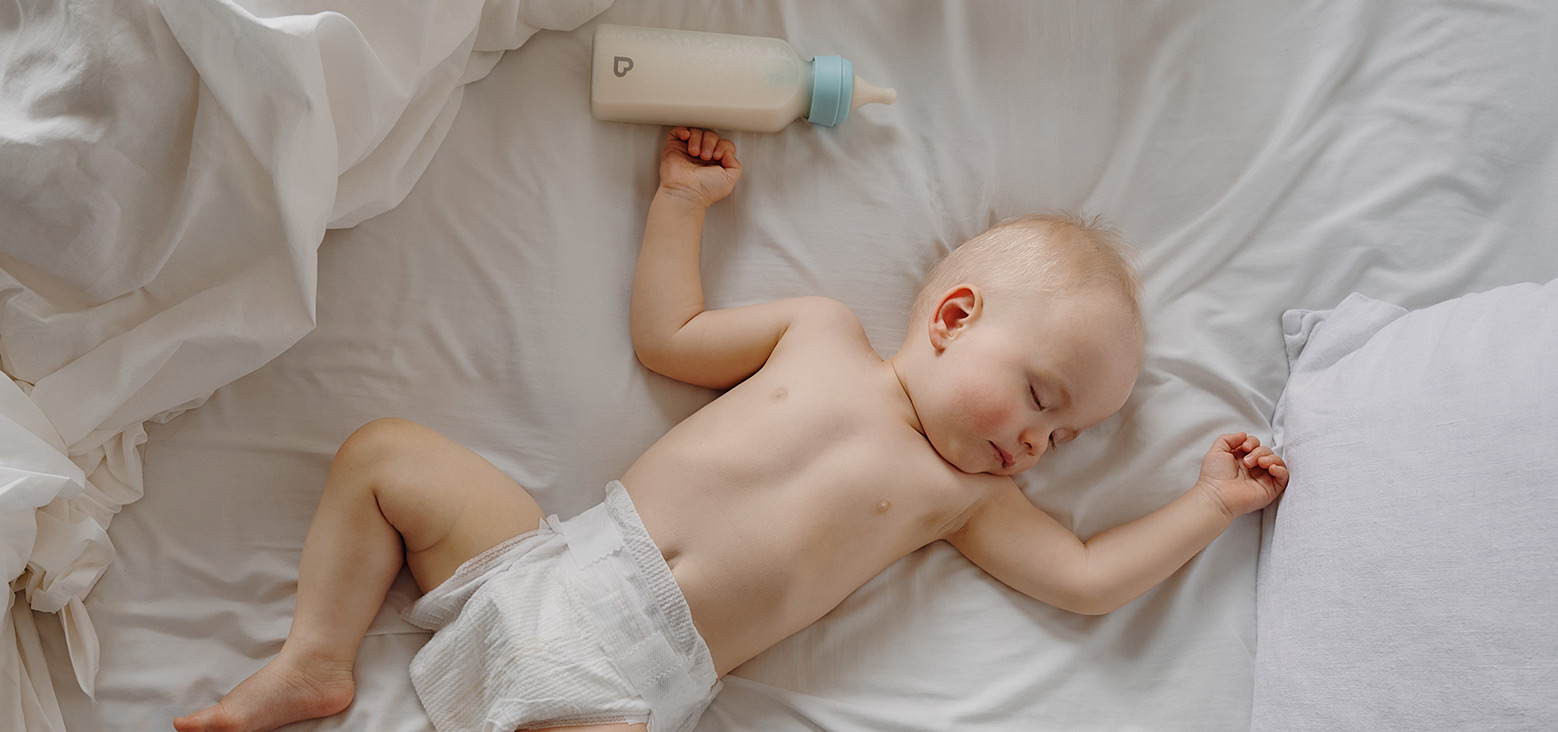 Baby sleeping on white bedding holding a Munchkin bottle with light blue band above their head.