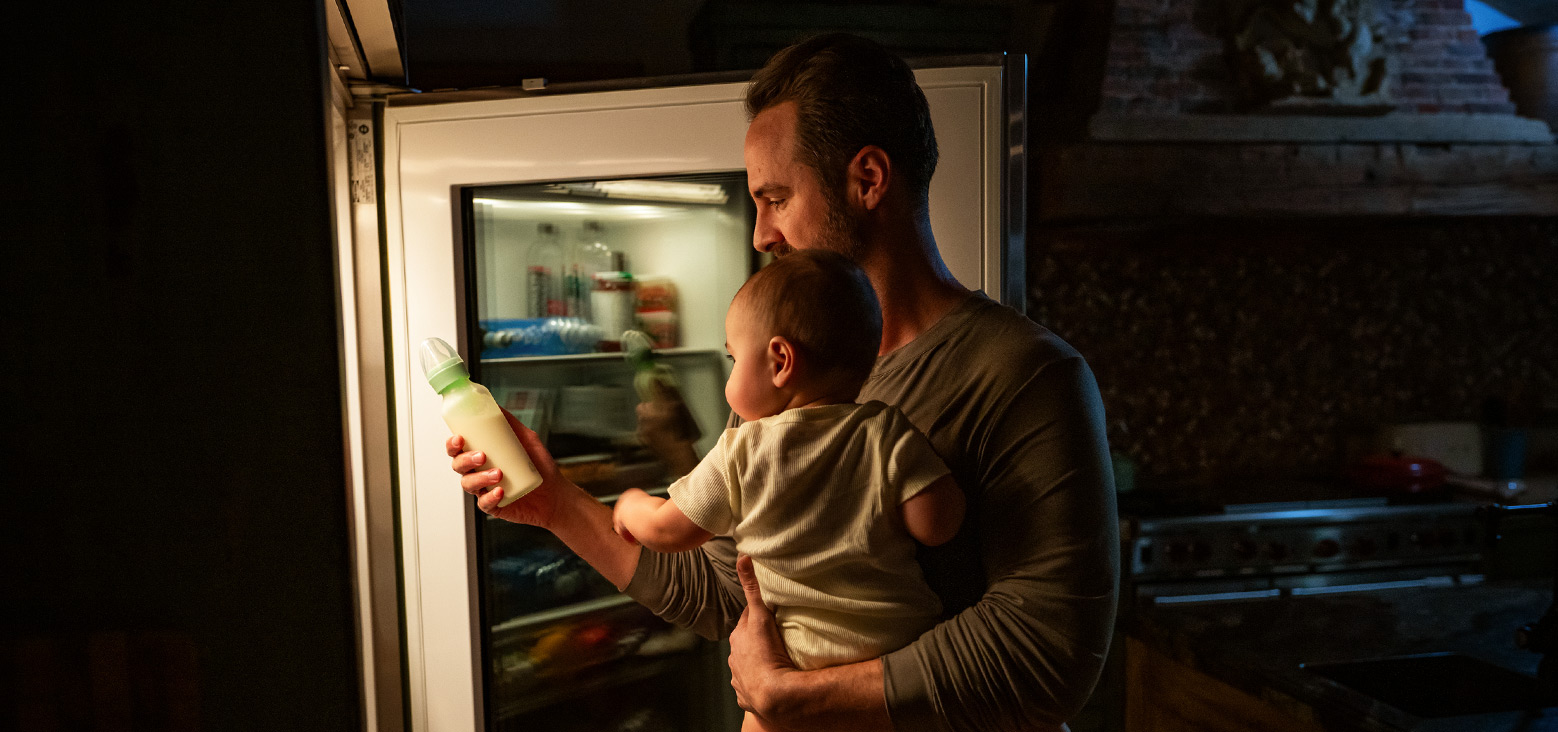 Father holding toddler while reaching into refrigerator to grab munchkin bottle