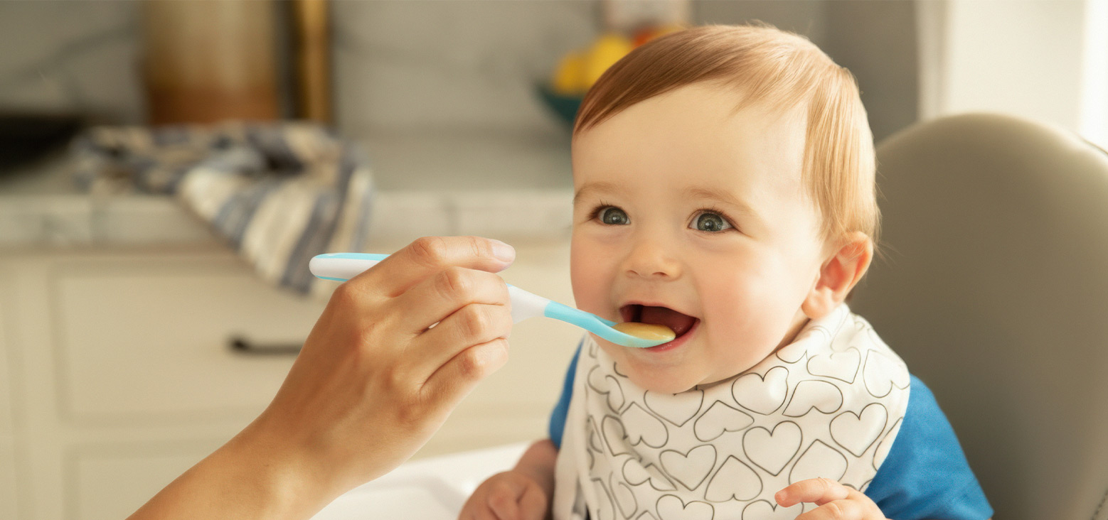 Toddler being fed with a blue and white spoon, wearing a heart-patterned bib, smiling in a bright kitchen.
