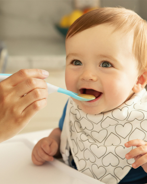 Happy baby eating from a blue and green spoon held by an adult's hand, wearing a heart-patterned bib.