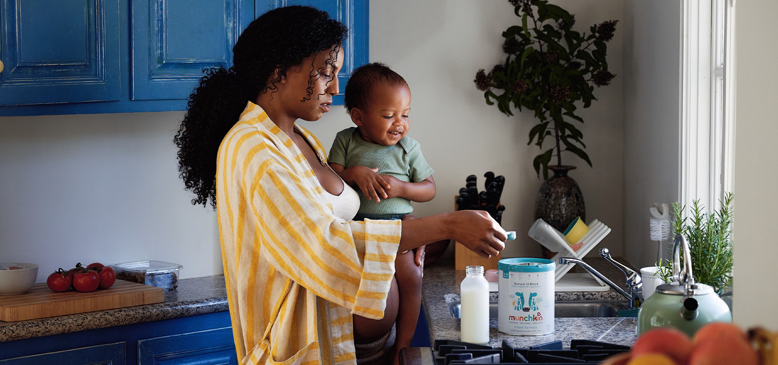 Woman in yellow striped shirt holding smiling toddler in kitchen, reaching for Munchkin formula container on counter next to milk bottle and kitchen items.