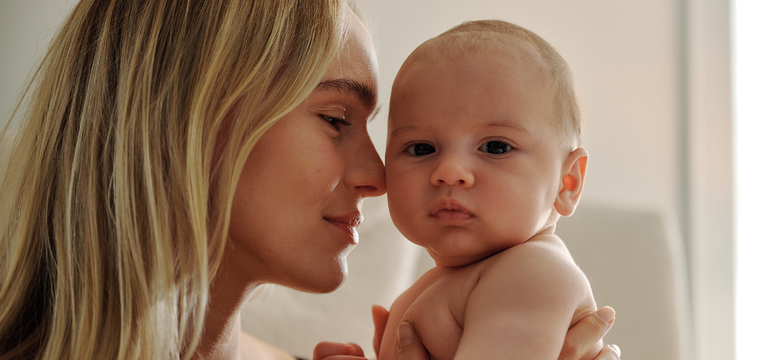 Mother with blonde hair holding and looking at her infant baby in a bright, warm setting.
