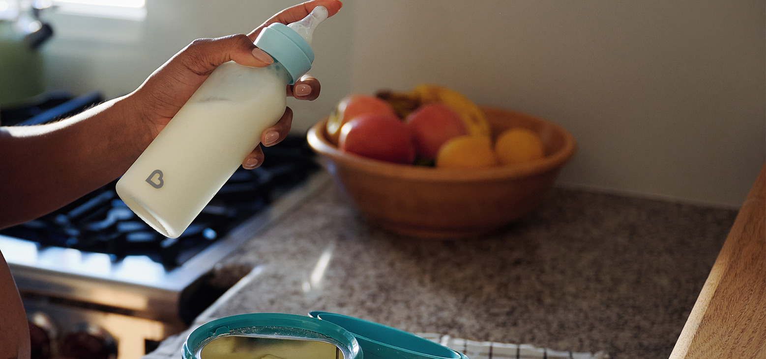 Adult hand holding a Munchkin bottle with blue cap above a bowl of food, with a fruit bowl blurred in the background.