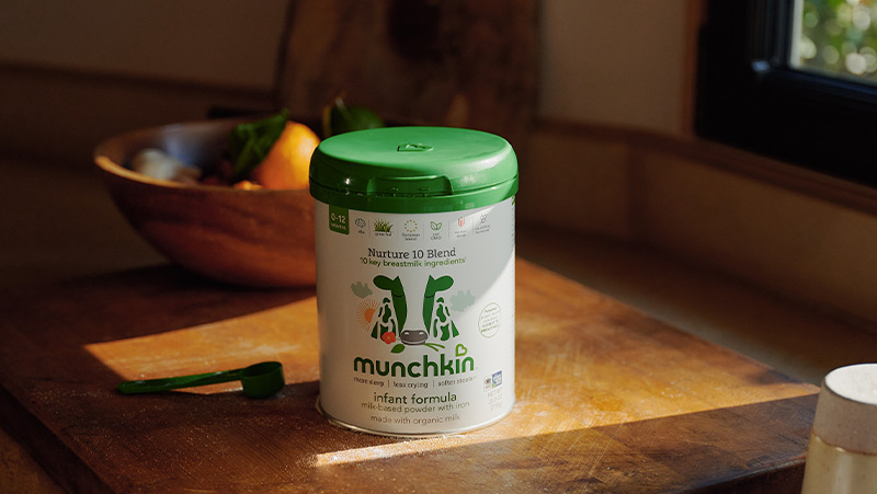 Munchkin Nurture 10 Blend infant formula container with green lid on a wooden table in a home kitchen setting.