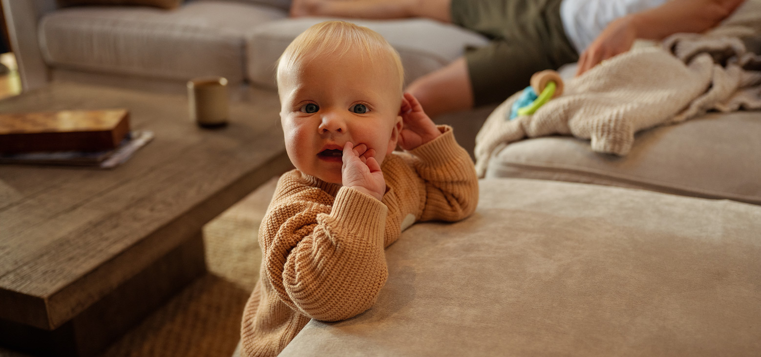 Baby in a tan knit sweater playing on a living room floor while caregivers supervise nearby.