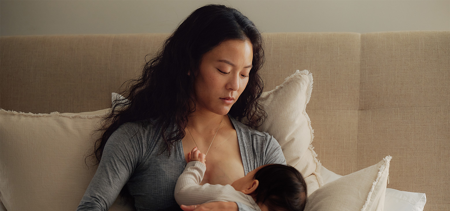 Mother nursing infant on a beige couch with cream pillows in a calm, intimate moment.