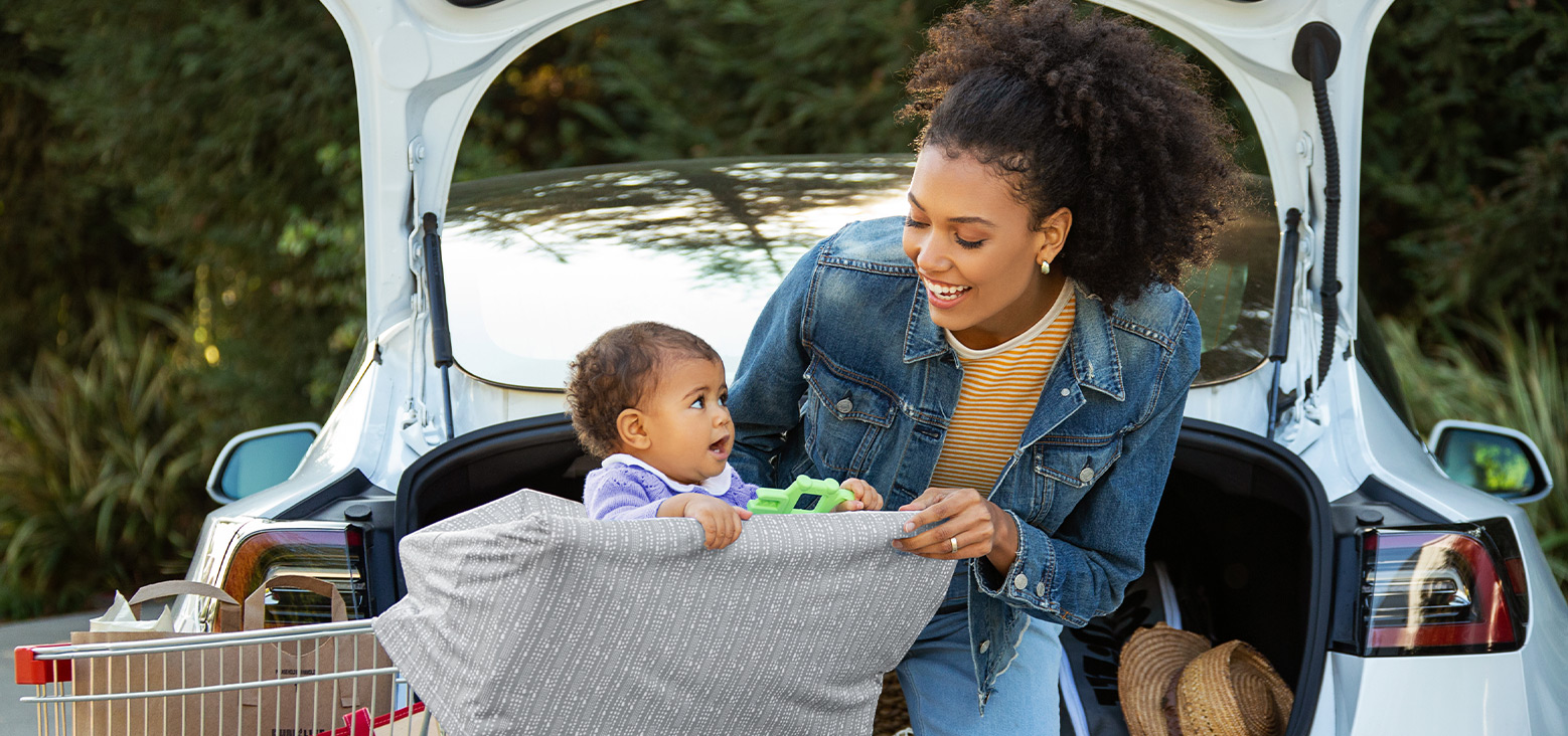 Parent and toddler loading a car trunk with groceries and a Munchkin product, outdoor setting with green foliage in background.