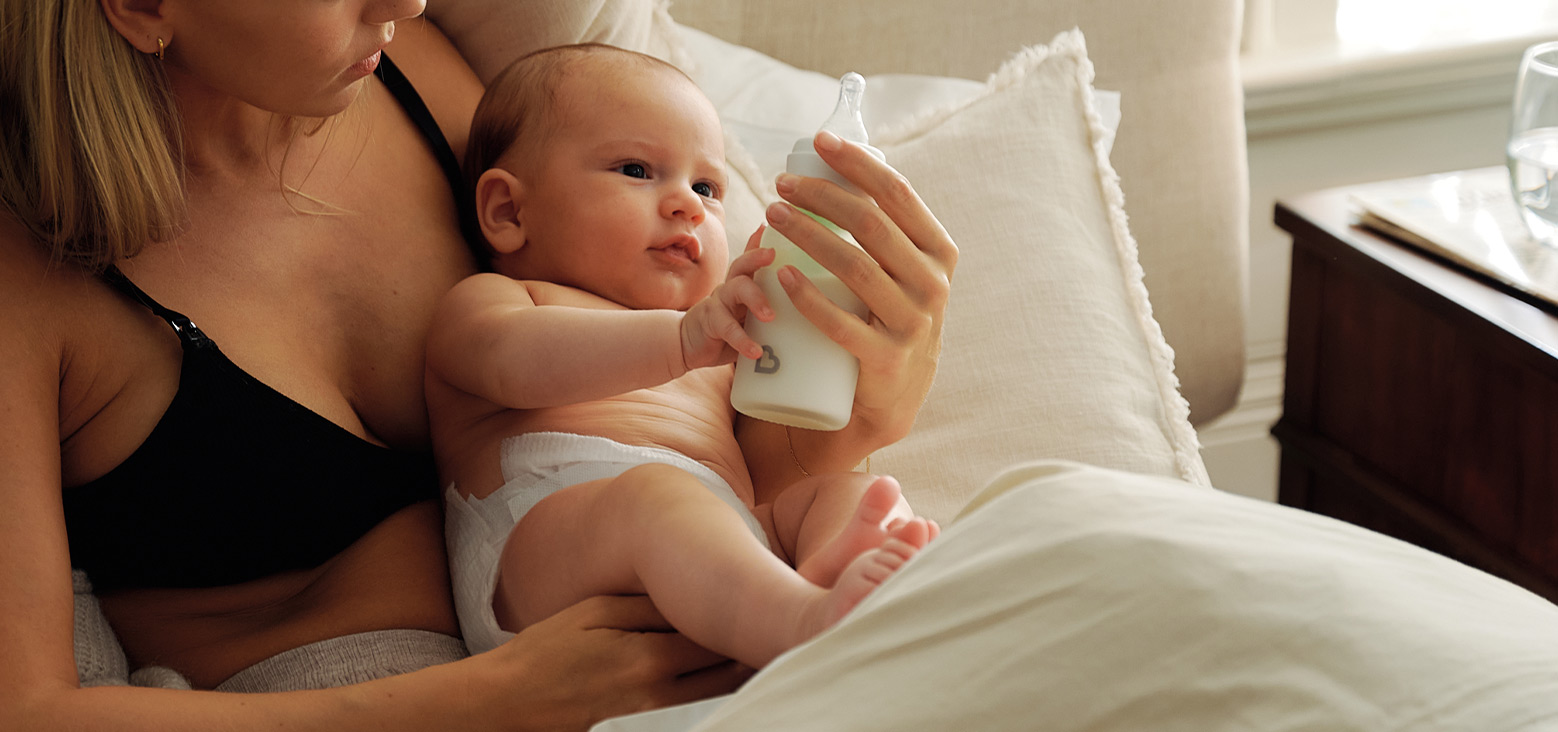 Parent holding infant on bed while baby feeds from bottle