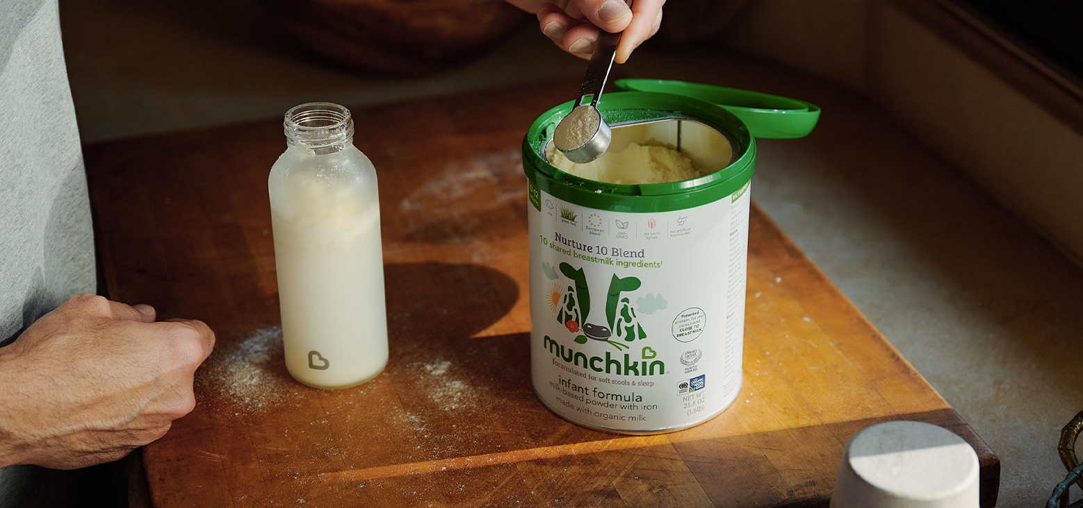 Adult hand holding Munchkin Nurture 10 Blend infant formula container with green lid and scoop, next to bottle of formula to be mixed on wooden surface.