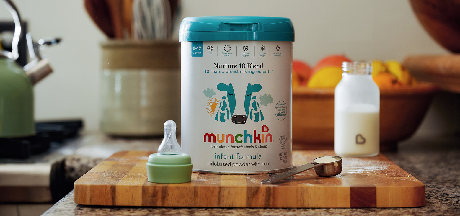 Munchkin Nurture 10 Blend infant formula canister with teal lid on wooden cutting board, surrounded by baby bottle and feeding supplies in kitchen setting.