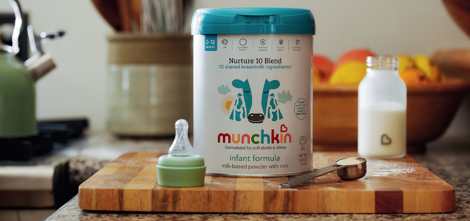 Munchkin Nurture 10 Blend infant formula canister with turquoise lid displayed on a wooden cutting board alongside a green bottle with nipple and measuring scoop in a kitchen setting.