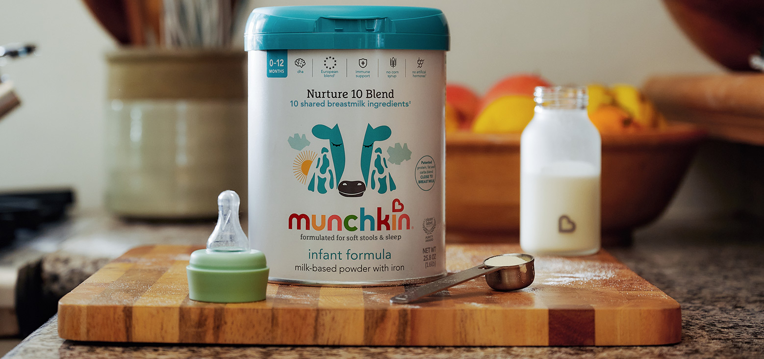 Munchkin Nurture 10 Blend infant formula container with teal lid on a wooden cutting board, alongside a green bottle nipple and measuring spoon, with a bottle of prepared formula in the background on a kitchen counter.