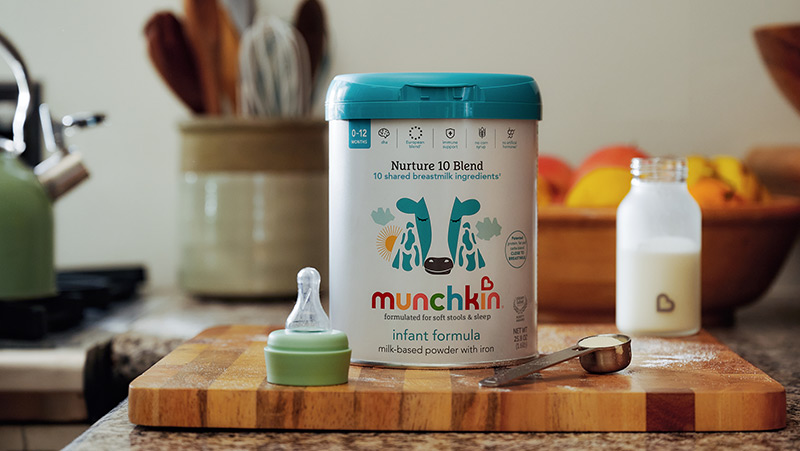 Munchkin Nurture 10 Blend infant formula container with bottle and nipple on wooden surface in kitchen setting.