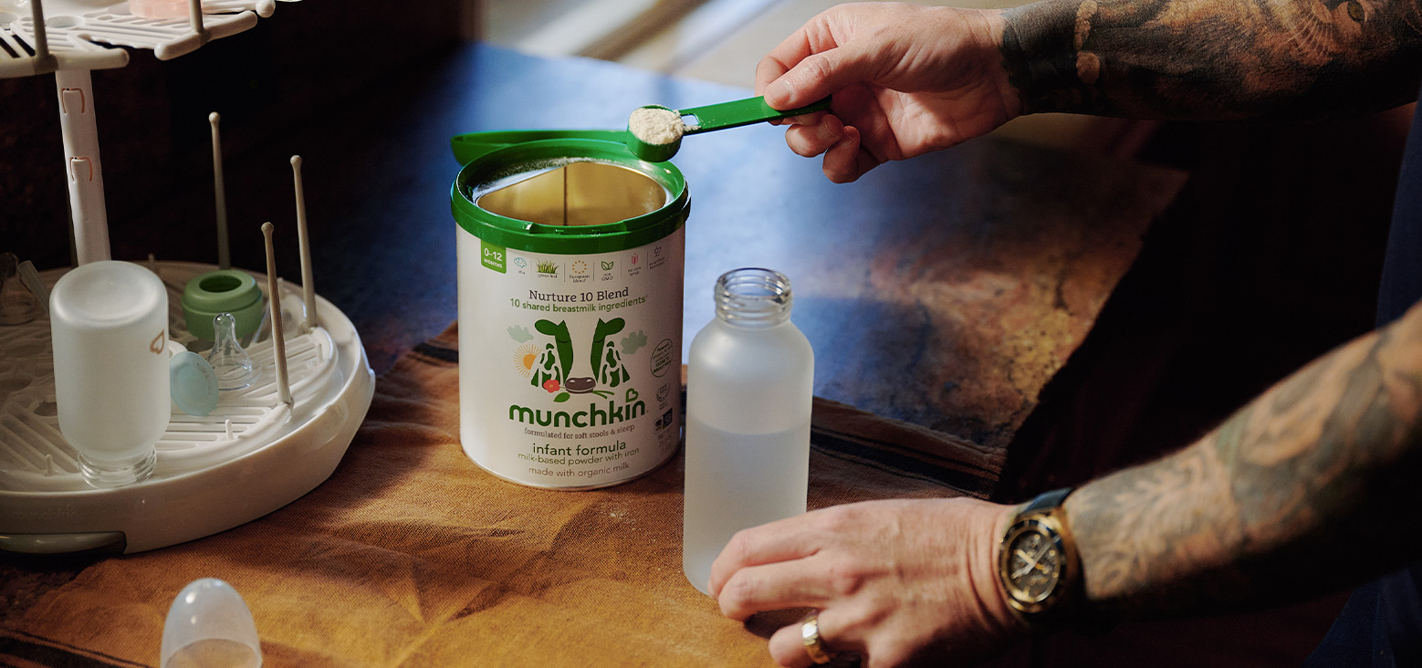 Adult using a green Munchkin scoop to prepare Nurture 10 Blend infant formula in a bottle, with formula preparation station and accessories visible on wooden table.