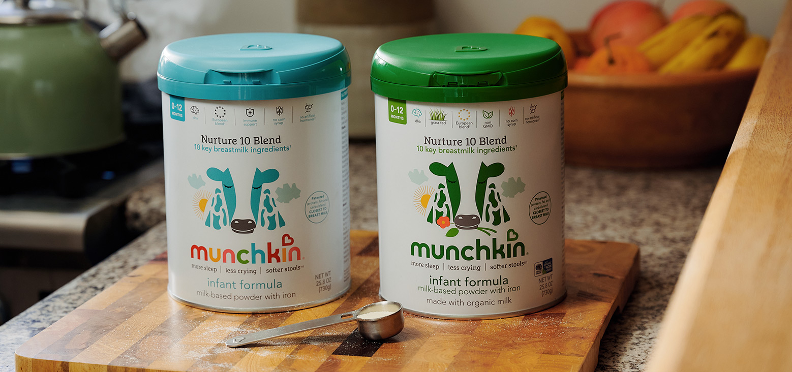 Two Munchkin 10 Blend infant formula cans with turquoise and green lids on a wooden surface in a kitchen, with a measuring scoop nearby and fruit in the background.