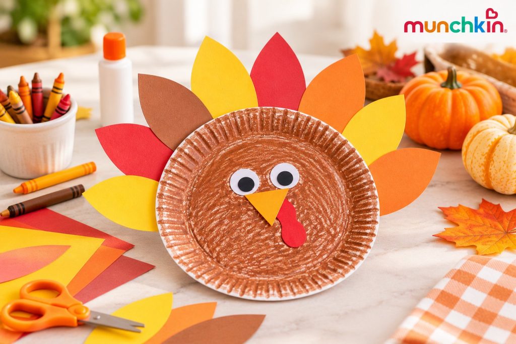 Munchkin Thanksgiving paper-plate turkey craft featuring a brown crayon-coloured paper plate decorated with googly eyes, a paper beak, red wattle, and colourful red, orange, yellow, and brown paper feathers, surrounded by crayons, craft supplies, and autumn decorations.