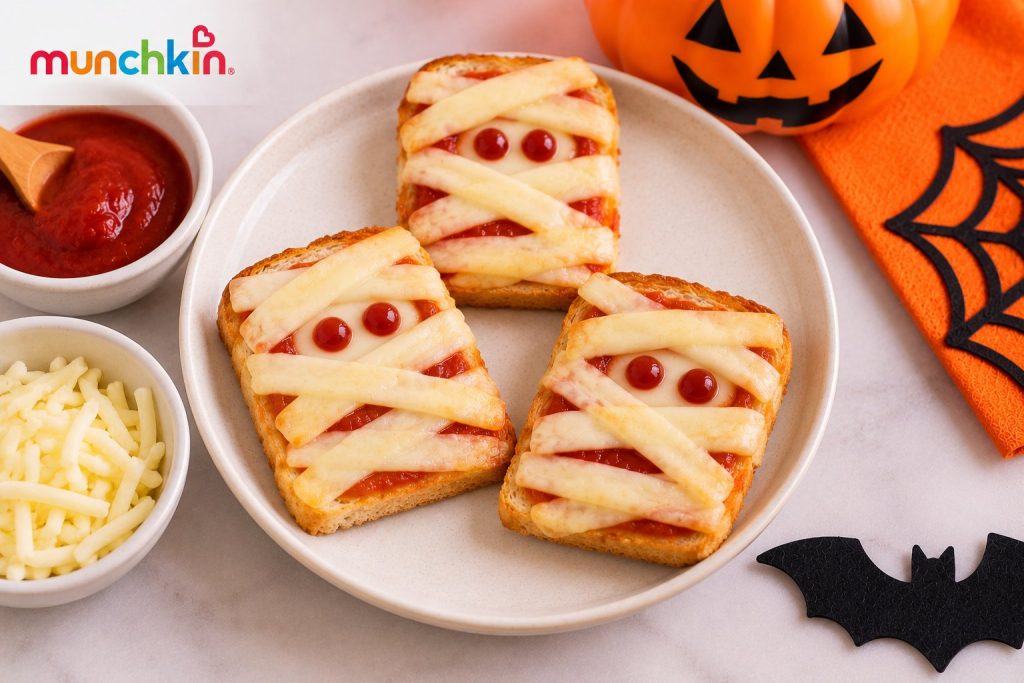 Munchkin Halloween mini mummy toast made with bread, tomato sauce and thin cheese strips arranged like mummy bandages, with small sauce-dot eyes, served on a plate alongside shredded cheese and tomato sauce with festive Halloween decorations.