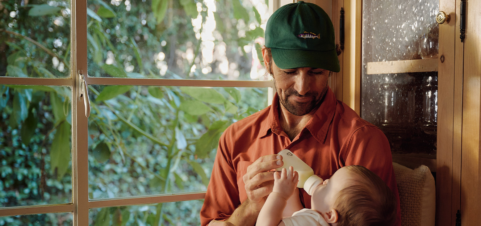 Adult holding and feeding a baby with a Munchkin bottle by a window with green foliage in the background.