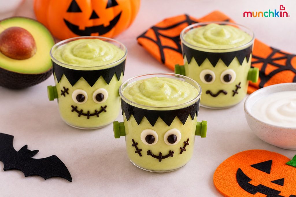 Munchkin Halloween snack featuring green avocado-yogurt cups decorated as Frankenstein faces, with playful eyes, stitched smiles, and black paper hair, surrounded by avocado and festive Halloween decorations.