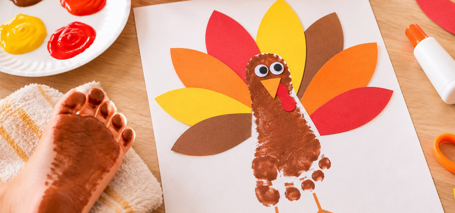 Toddler's footprint art project featuring a turkey made from a brown handprint, colorful paper feathers, googly eyes, and an orange beak, surrounded by paint supplies and craft materials on a wooden surface.