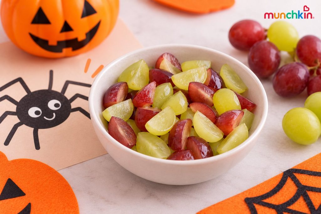 Spider-egg grapes made with quartered red and green grapes in a white bowl, styled as a Halloween snack with a friendly spider drawing and festive pumpkin decorations.
